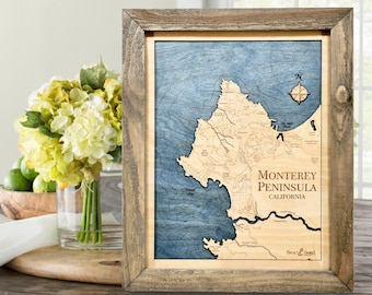 May include: Wooden framed map of Monterey Peninsula, California, with a blue ocean and tan landmass. The map includes detailed topography and the text "Monterey Peninsula California". A compass rose is in the upper right corner.