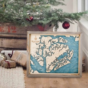 May include: Framed wooden map of Beaufort, North Carolina, with a detailed depiction of the coastline and waterways in shades of blue and beige. The map is encased in a gray wooden frame and features a compass rose and the city name.