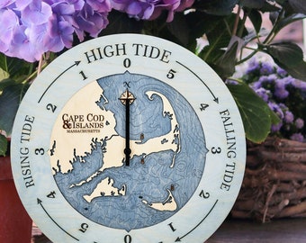 May include: A wooden tide clock with a blue and white map of Cape Cod and the Islands, Massachusetts. The clock face has a black hand that points to the current tide level. The clock face is labeled "High Tide", "Low Tide", "Rising Tide", and "Falling Tide".