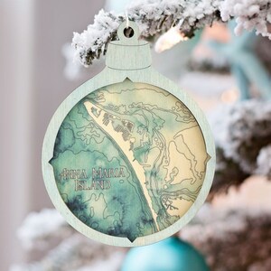 May include: A wooden Christmas ornament shaped like a bauble with a light blue background and a map of Anna Maria Island, Florida. The map is in shades of green and brown with the text "Anna Maria Island" printed in white.