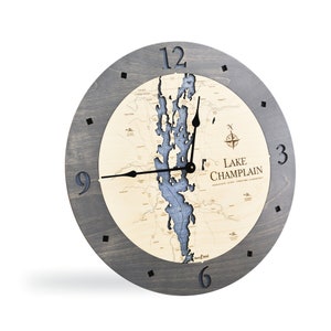 May include: A gray wooden wall clock with a map of Lake Champlain in the center. The clock face is surrounded by a gray wooden frame with black numbers and hands. The map is in shades of blue and brown, with the lake highlighted in blue. The clock has a compass rose in the lower right corner.