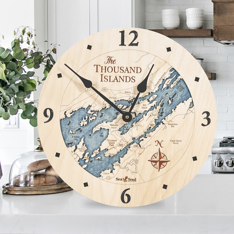 Puede incluir: Un reloj de madera con un mapa de las Mil Islas en la esfera. El reloj tiene agujas negras y un dise&ntilde;o de br&uacute;jula en la esquina inferior derecha. El texto "The Thousand Islands" est&aacute; impreso en marr&oacute;n en la esfera del reloj.