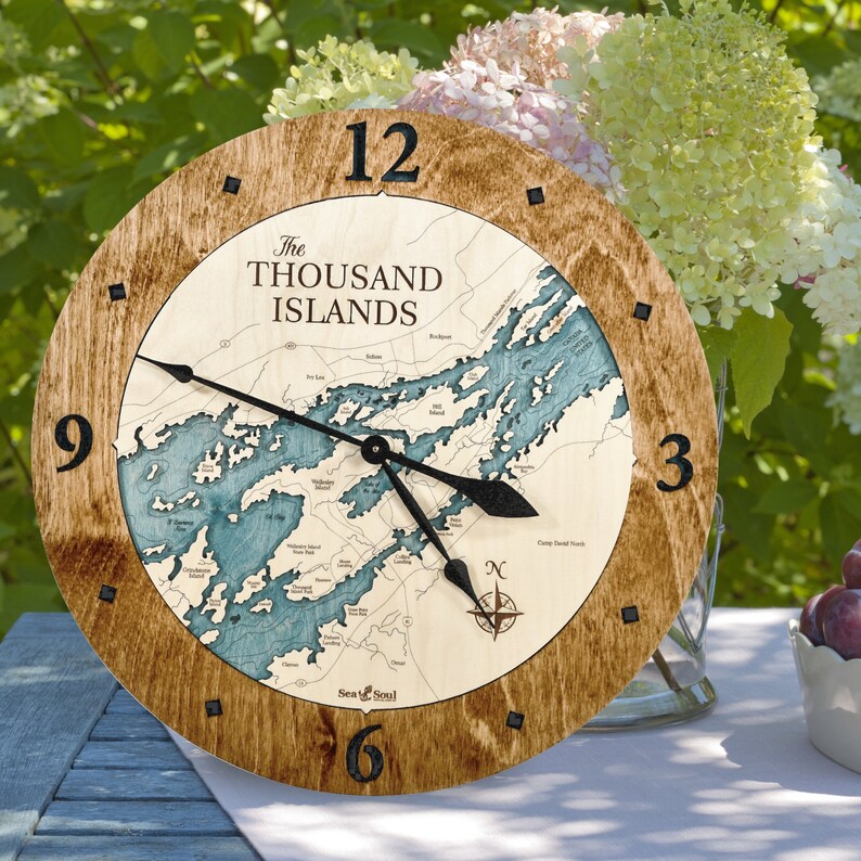 Puede incluir: Un reloj de madera con un mapa de las Mil Islas en la esfera. El reloj tiene agujas negras y un dise&ntilde;o de br&uacute;jula en el centro. El reloj est&aacute; rodeado por un marco de madera marr&oacute;n.