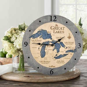 May include: A gray wooden clock with a map of the Great Lakes on the face. The clock face is surrounded by a gray wooden frame. The clock hands are black. The clock face includes the names of the Great Lakes: Superior, Huron, Ontario, Michigan, and Erie. The clock face also includes the names of the states and provinces that border the Great Lakes. The clock face also includes a compass rose.