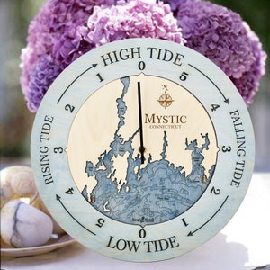 May include: A round wooden tide clock with a light blue outer ring and a natural wood center. The clock face features a map of Mystic, Connecticut, with text indicating high and low tide, and a compass rose.