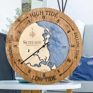 May include: A round wooden tide clock featuring a map of Scituate, Massachusetts. The face displays a light wood landmass and blue water with depth contours, a compass rose, and black clock hands. The outer ring shows tide levels and text.