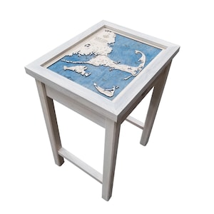 May include: A white wooden end table with a framed nautical map of Cape Cod, Massachusetts. The map is blue and white and shows the coastline and islands of the area.