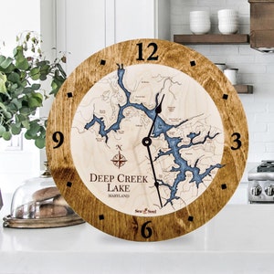 May include: A wooden clock with a map of Deep Creek Lake, Maryland. The clock face is brown with black numbers and a compass rose in the center. The map is blue and white and shows the lake's shoreline and islands. The clock is 12 inches in diameter.