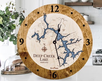 May include: A wooden clock with a map of Deep Creek Lake, Maryland. The clock face is brown with black numbers and a compass rose in the center. The map is blue and white and shows the lake's shoreline and islands. The clock is 12 inches in diameter.