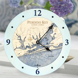 May include: A round wooden clock with a light blue frame and a detailed map of Perdido Key, Alabama-Florida. The clock face features black hands, black square markers, and large blue numerals. The text "Sea Soul" is at the bottom.