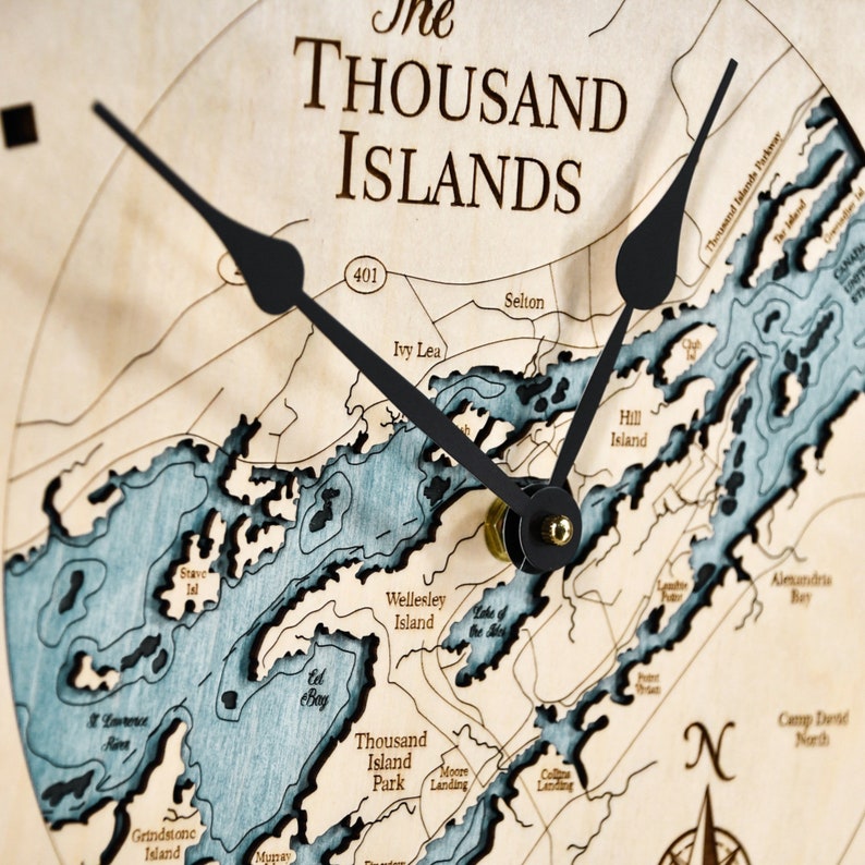 Puede incluir: Un reloj de madera con un mapa de las Mil Islas. La esfera del reloj tiene agujas negras y un mapa azul y blanco de las islas. La esfera del reloj tambi&eacute;n incluye el texto "The Thousand Islands" y "401".