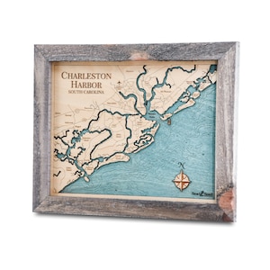 May include: A wooden map of Charleston Harbor, South Carolina, in a gray wood frame. The map is laser-cut and shows the coastline, waterways, and islands. The map is mounted on a blue background and includes a compass rose and the text "Sea & Soul".
