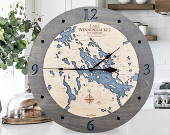 May include: A gray wooden clock with a map of Lake Winnipesaukee, New Hampshire. The clock face has black hands and black Roman numeral hour markers. The map is in shades of blue and brown.