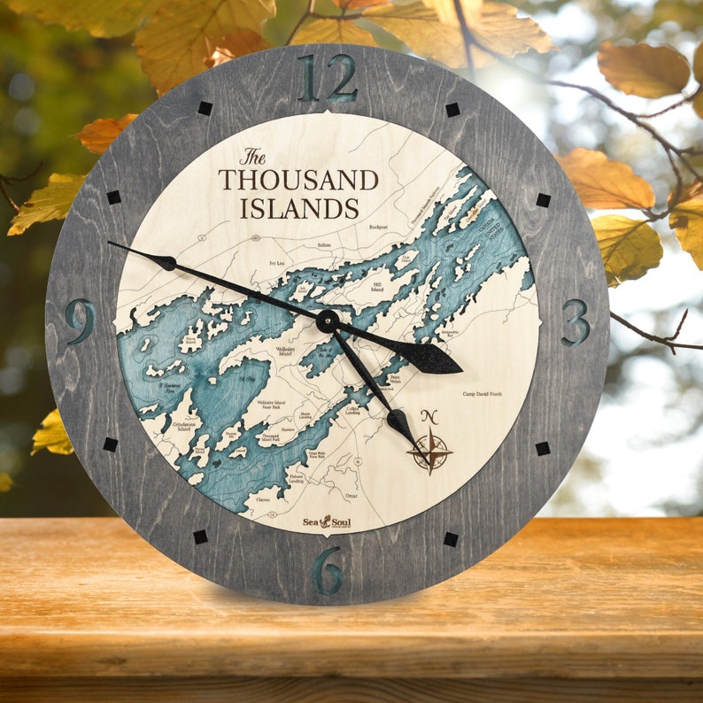 Puede incluir: Un reloj de madera con un marco de madera gris y un mapa de las Mil Islas en la esfera. Las manecillas del reloj son negras y los n&uacute;meros son de color gris oscuro. El reloj est&aacute; sobre una superficie de madera.