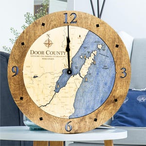May include: A round wooden clock with a map of Door County, Wisconsin. The clock face features a detailed map with blue water and tan land, with the text "Door County" and the time is approximately 12:00. The clock has black hands and black square markers.