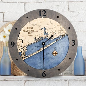 May include: A wooden clock with a map of East Matagorda Bay, Texas. The clock face is gray with black numbers and hands. The map is blue and brown with a compass rose in the center.