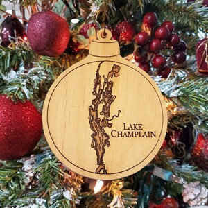 May include: A wooden Christmas ornament shaped like Lake Champlain, a large lake in the northeastern United States. The ornament is brown and has a detailed outline of the lake. The text "LAKE CHAMPLAIN" is engraved on the ornament.