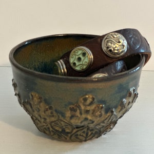 May include: A brown ceramic bowl with a leafy design and a brown leather bracelet with silver and green accents resting inside.