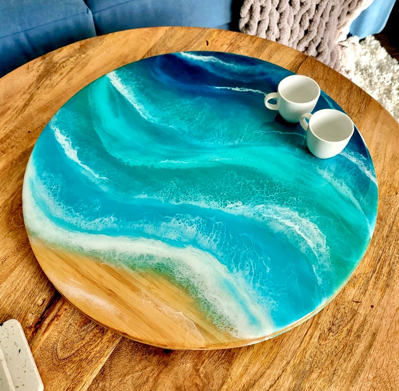 May include: A round wooden serving tray with a blue and white epoxy resin design that resembles ocean waves. The tray is on a wooden table with two white ceramic cups.