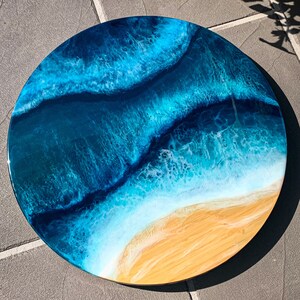 May include: A round resin art piece depicting an ocean scene with blue and white waves crashing on a sandy beach. The resin is clear and glossy, giving the piece a 3D effect.