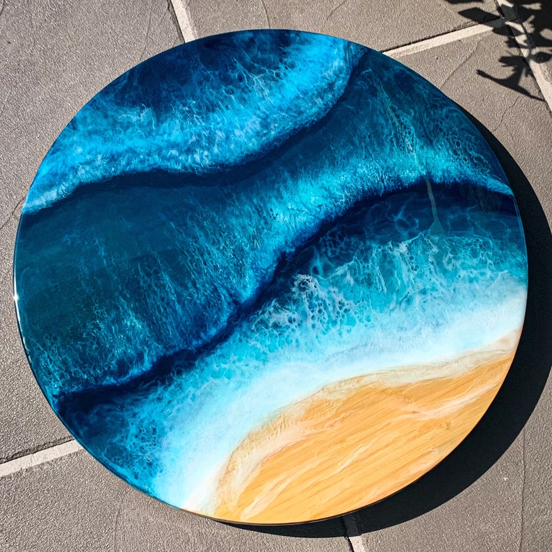 May include: A round resin art piece depicting an ocean scene with blue and white waves crashing on a sandy beach. The resin is clear and glossy, giving the piece a 3D effect.
