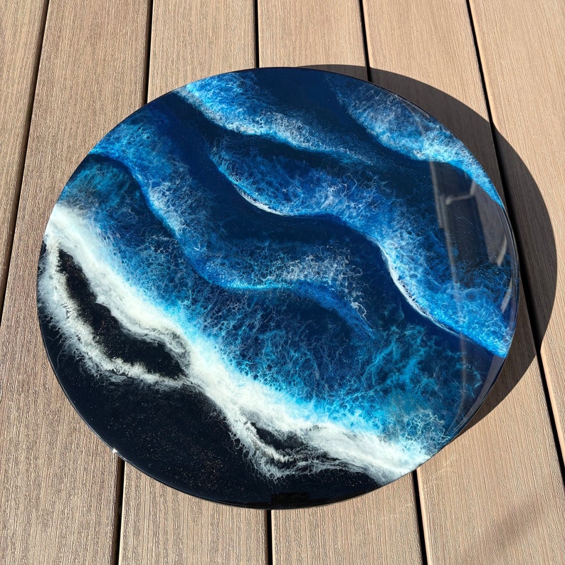 May include: A round resin art piece with a blue and white abstract design resembling ocean waves. The background is black with a subtle sparkle.