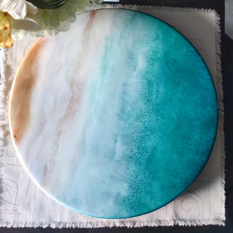 May include: Round resin art piece with a beach scene. The top half of the piece is white with brown accents, representing sand. The bottom half is a teal blue color, representing the ocean.