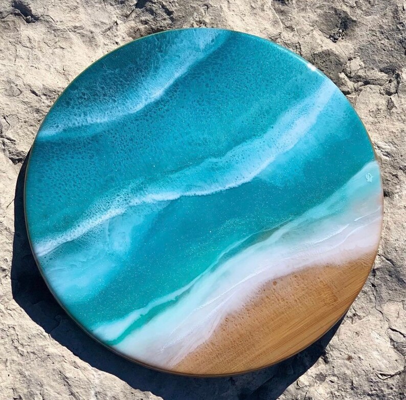 May include: Round wooden serving tray with a blue and white resin ocean wave design. The tray has a light brown wood border.