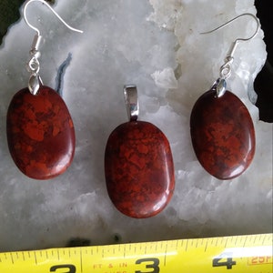 May include: A set of red jasper earrings and a pendant. The stones are oval shaped and have a mottled pattern. The earrings are silver and have a hook closure. The pendant is silver and has a bail.