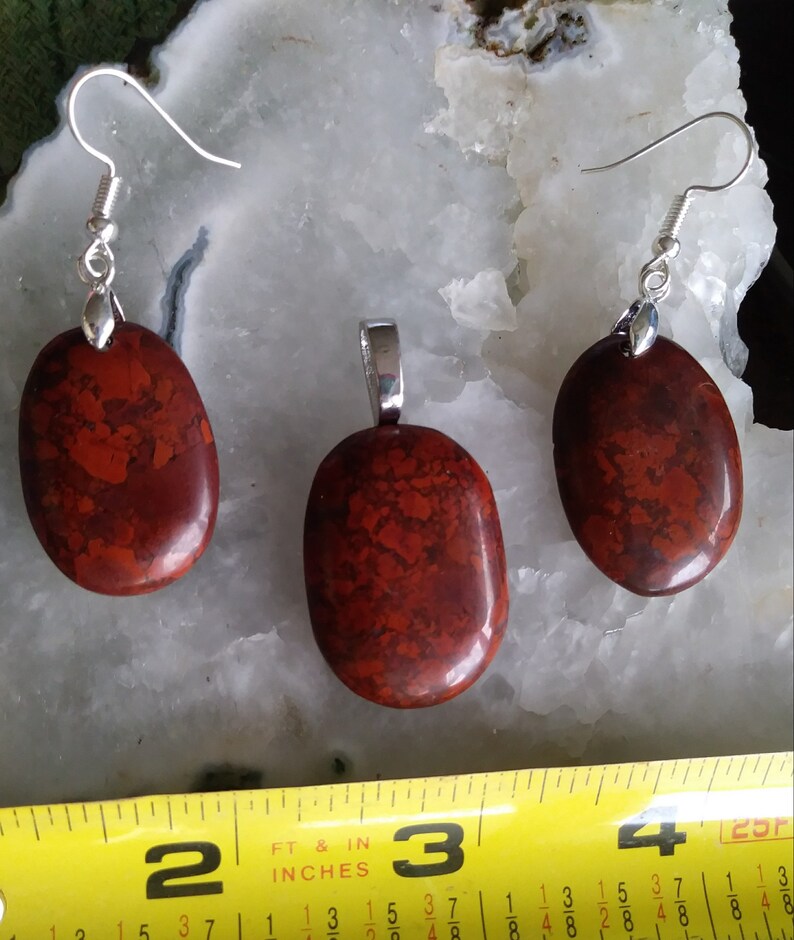 May include: A set of red jasper earrings and a pendant. The stones are oval shaped and have a mottled pattern. The earrings are silver and have a hook closure. The pendant is silver and has a bail.