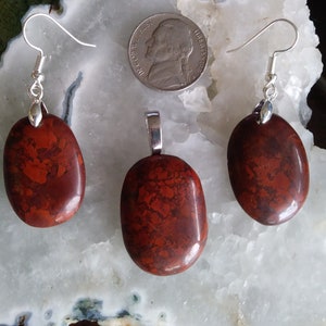 May include: A set of red jasper jewelry featuring a pendant and earrings. The stones are a mottled red and brown color with a smooth, oval shape. The pendant and earrings are attached to silver-toned chains.