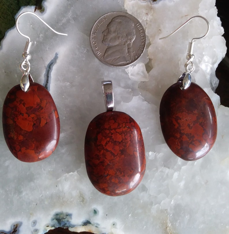 May include: A set of red jasper jewelry featuring a pendant and earrings. The stones are a mottled red and brown color with a smooth, oval shape. The pendant and earrings are attached to silver-toned chains.