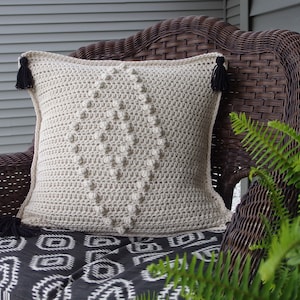 May include: A cream-coloured crocheted cushion with black tassels sits on a wicker chair with a black and white patterned cushion. A green fern plant is in the background.