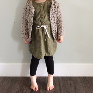May include: A young person wearing a beige crocheted cardigan over a green dress with a white drawstring. The person is wearing black leggings and has bare feet.