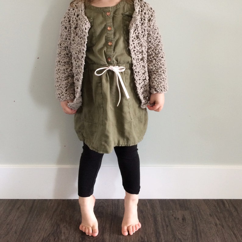 May include: A young person wearing a beige crocheted cardigan over a green dress with a white drawstring. The person is wearing black leggings and has bare feet.