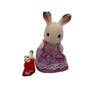 May include: Two toy rabbits. One is larger and wears a pink dress with white polka dots and lace trim. The smaller rabbit is wrapped in a red blanket. Both rabbits have white fur with brown ears.