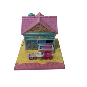 Op de afbeelding: Een miniatuur café speelset met een roze dak en het woord "CAFE" op de luifel. Het gebouw heeft blauwe en groene accenten, met een kleine tafel en stoelen buiten. De basis is geel en roze.