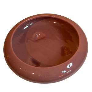 May include: A round, terracotta-colored ceramic bowl with a smooth, glossy finish. The interior features three small, raised, rounded protrusions integrated into the base, creating a distinct texture within the shallow, circular basin.