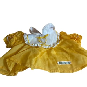 May include: A bright yellow dress with short puffed sleeves and a white collar featuring a subtle vertical stripe pattern. The collar is accented with a white eyelet lace trim and a small yellow ribbon tie at the center. A small rectangular label is visible.