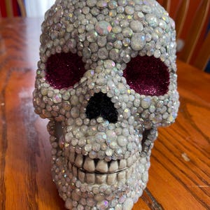 May include: A decorative skull covered in iridescent rhinestones. The eye sockets are filled with sparkly magenta material, and the nose is black. The skull is a unique, eye-catching piece.