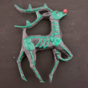 以下が含まれることがあります： 黒と緑の骨格デザインのトナカイのオーナメント。トナカイは赤い鼻と緑の角をしています。