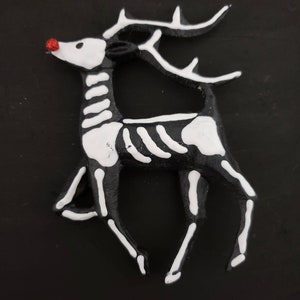以下が含まれることがあります： 赤い鼻をした黒と白の骨格のトナカイのオーナメント。トナカイは後ろ足で立ち、前足を上げています。