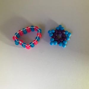May include: A beaded bracelet and a star-shaped charm. The bracelet is made of pink and blue beads strung together with white string. The star charm is made of blue and purple beads. Both items are on a white surface.