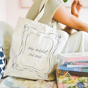 May include: Cream-colored tote bag with a black frame design and the text "Mrs. Mitchell 2nd Grade." The bag has a shoulder strap and is sitting next to a stack of books with floral and pastel covers. A black and white cow print pillow is visible.