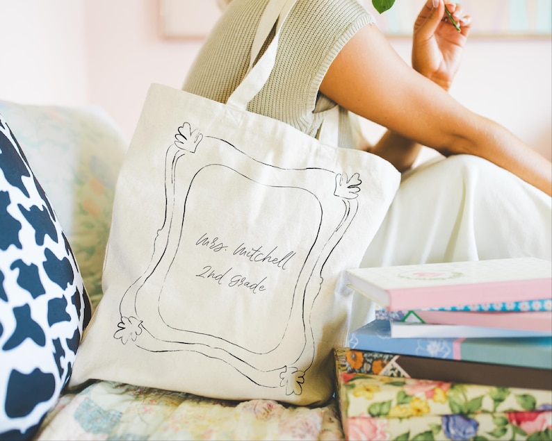 May include: Cream-colored tote bag with a black frame design and the text "Mrs. Mitchell 2nd Grade." The bag has a shoulder strap and is sitting next to a stack of books with floral and pastel covers. A black and white cow print pillow is visible.