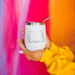 May include: White insulated tumbler with a silver rim and straw, featuring a black line drawing of an open book. The tumbler is held in hands, against a colorful pink, blue, and orange background. A yellow sleeve is visible.