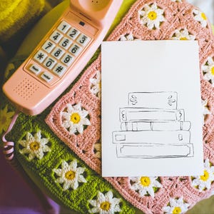 May include: A vintage pink telephone rests on a crocheted blanket with daisy patterns in pink, green, and white. A white card with a black line drawing of stacked books is next to the phone. The scene has a retro, cozy aesthetic.