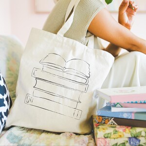 May include: Cream-colored tote bag with a black line drawing of a stack of books. The bag has long straps and is sitting next to a stack of books with colorful covers. The image has a soft, natural light.