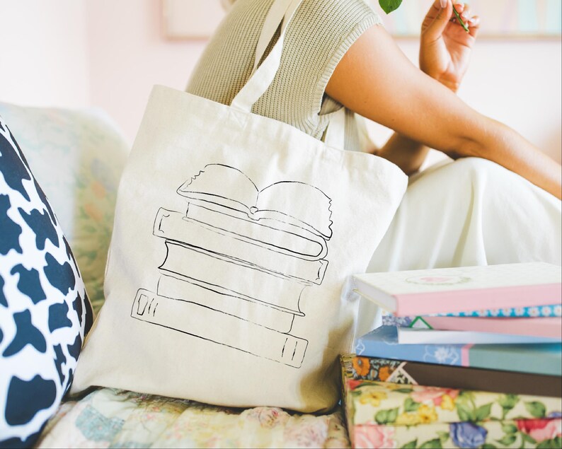 May include: Cream-colored tote bag with a black line drawing of a stack of books. The bag has long straps and is sitting next to a stack of books with colorful covers. The image has a soft, natural light.