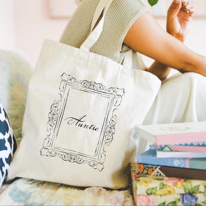 May include: Cream-colored tote bag with a black ornate frame design and the word "Auntie" printed inside. The bag has a simple strap and is sitting next to a stack of books with floral and pastel covers.